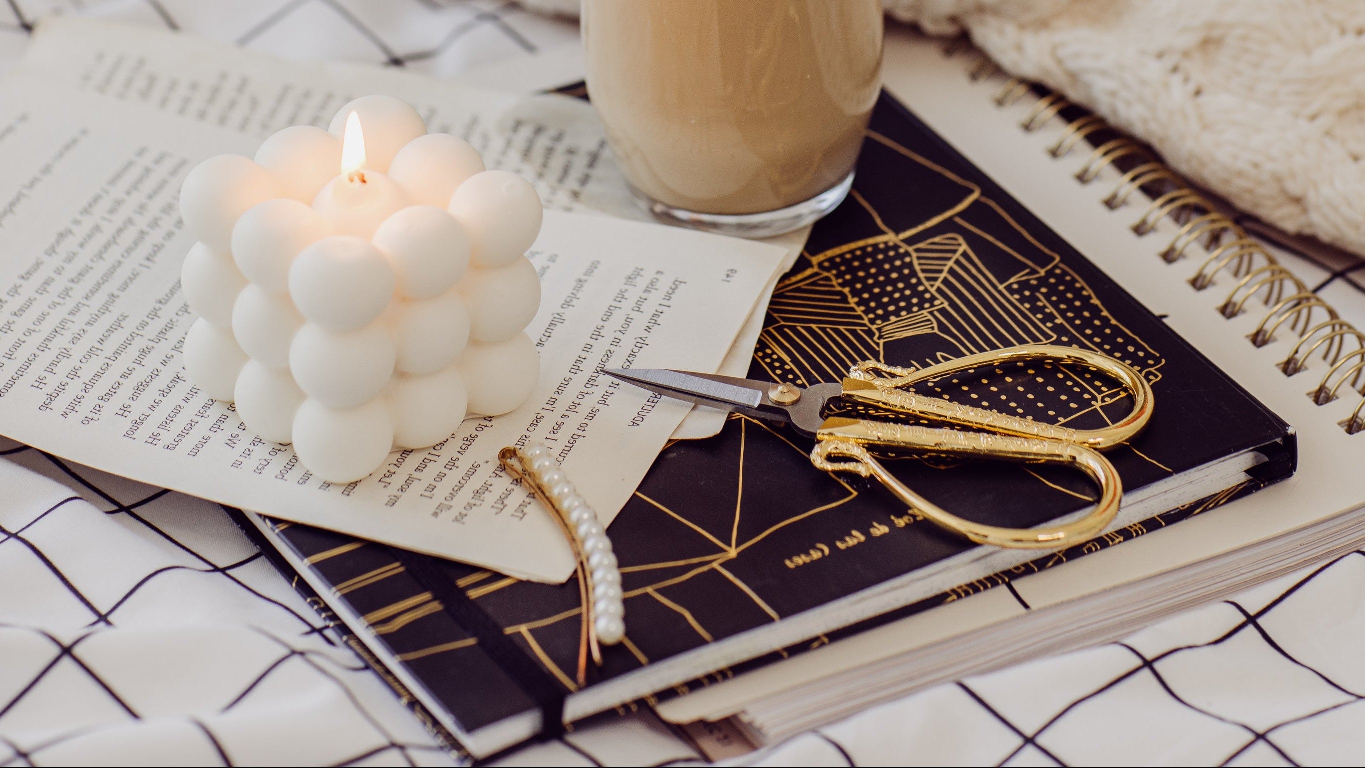 Notebook with gold scissors, a glass of coffee, and a white candle on a checkered tablecloth.